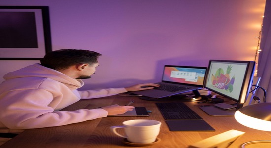 Designer using laptop and tablet at desk with dual monitors for digital artwork.
