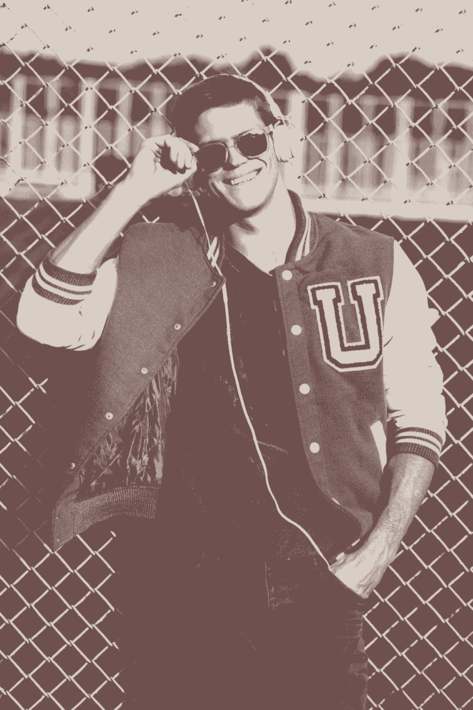 Smiling young man in a varsity jacket with letter patch, listening to music by a fence.