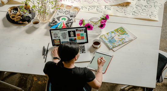 Designer working with laptop, tablet, and sketches at a creative workspace.