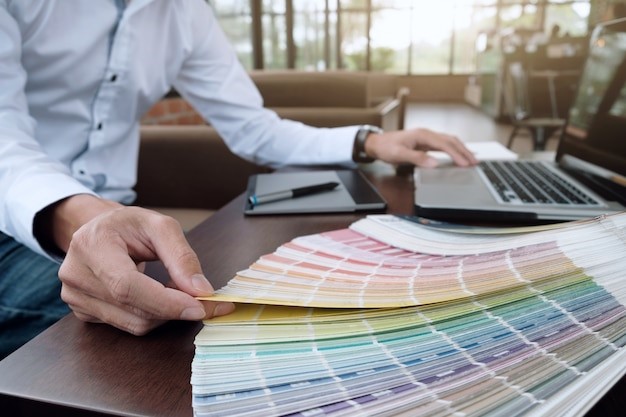 Designer reviewing a color palette fan deck beside a laptop on a desk.