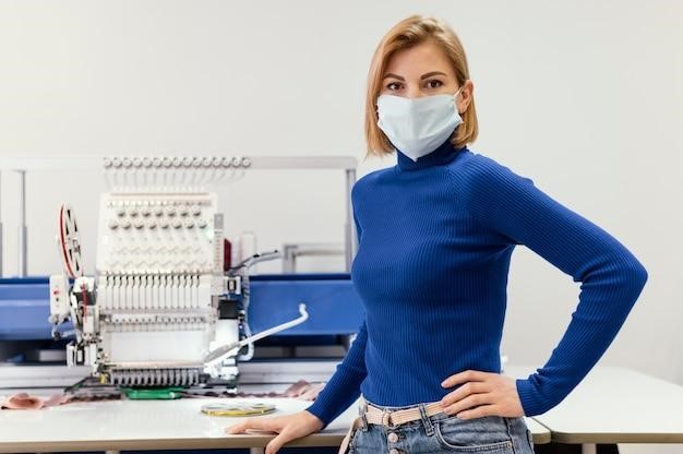 Woman in mask standing beside embroidery machine in workshop.