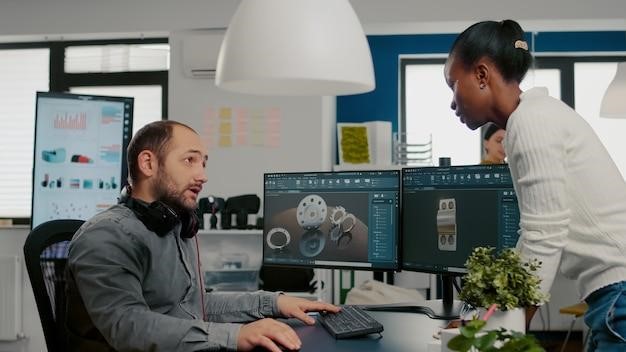 Designer discussing 3D models on dual monitors with a colleague in office.