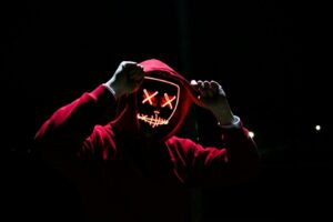 Person in red hoodie with glowing mask against a dark background.