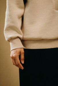 Person wearing a beige sweatshirt and black pants standing against a plain background.