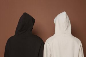 Two hooded sweatshirts — one black and one cream — shown from the back against a warm brown backdrop.