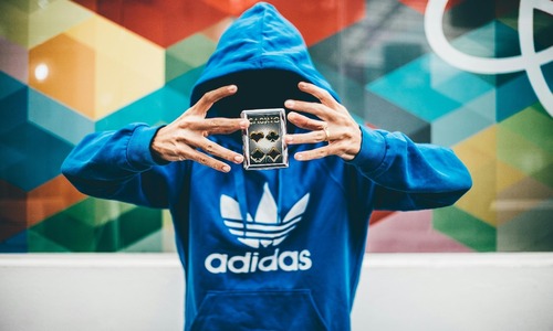 Man in blue Adidas hoodie performing a card magic trick with vibrant geometric background.