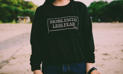 White sweater with “More Faith Less Fear” text printed on the front, minimalist flat lay apparel.