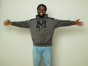 Smiling man with arms outstretched wearing a grey University of Michigan hoodie