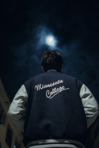 Back view of a person wearing a varsity jacket reading "Minnesota College" under a moonlit night sky.