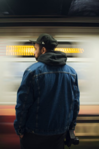 Rear view of a man wearing a denim jacket, black hoodie, and cap in front of a moving train.